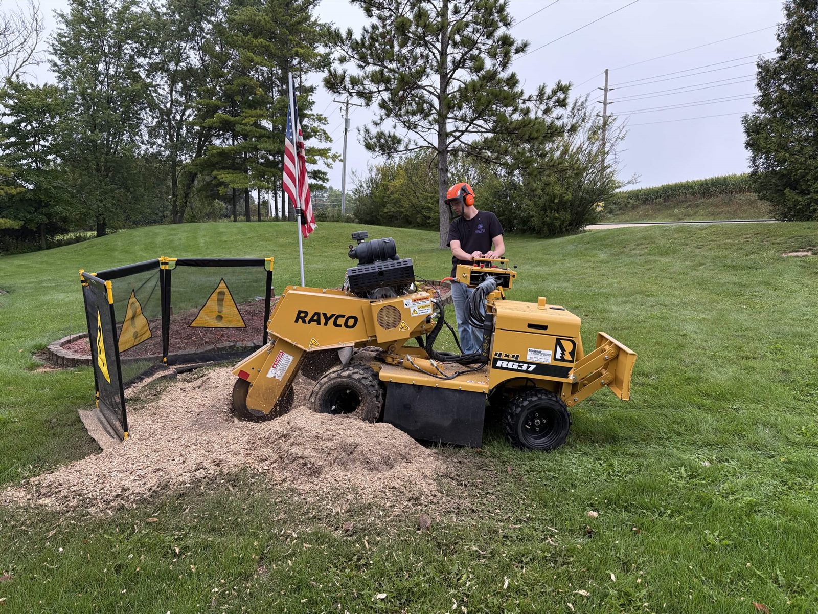 Austin at the controls of the Rayco RG37, safety screens up, grinding a stump beside a flower bed